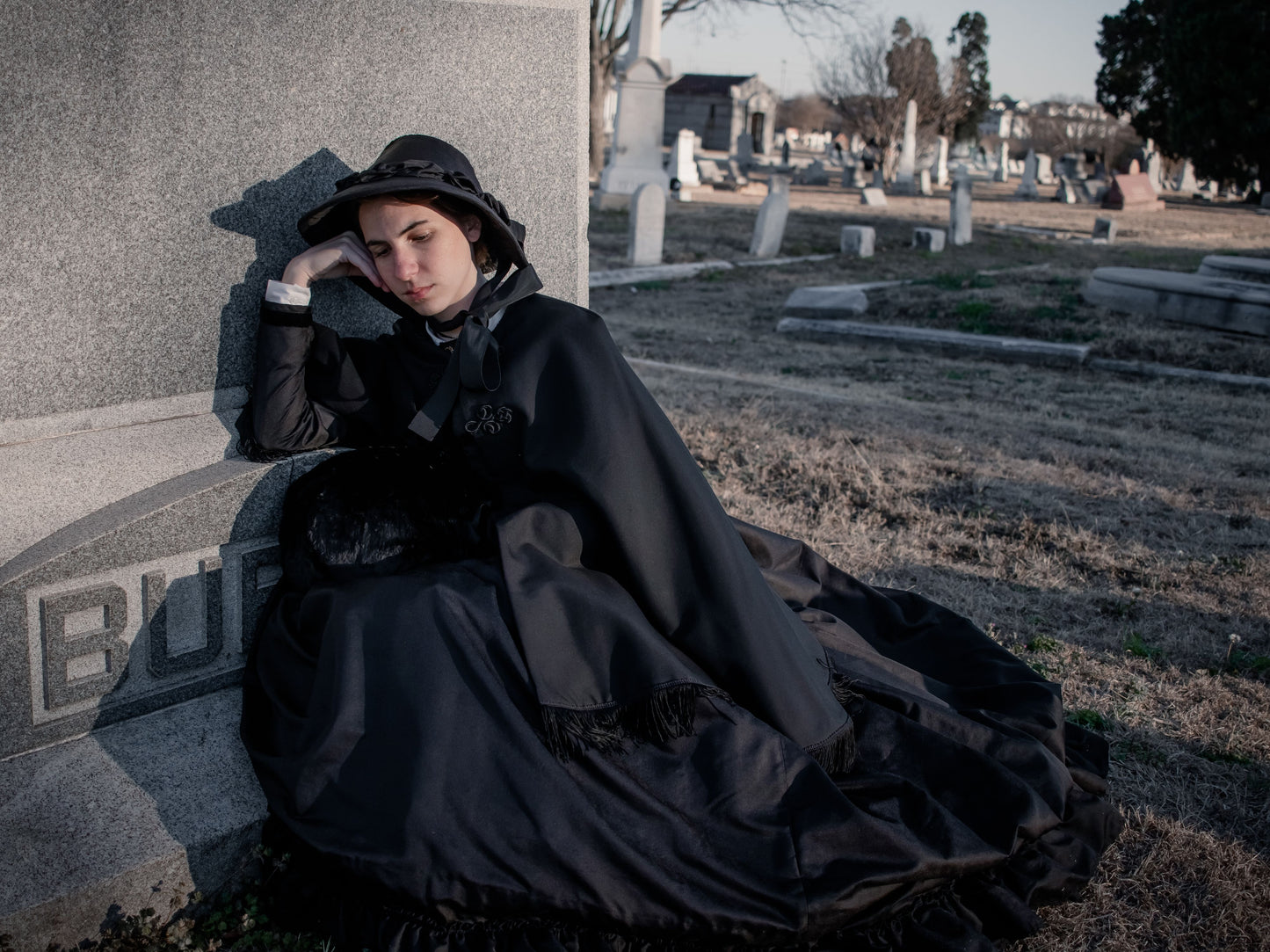 Model sits on dead brown grass and leans against a large headstone. She wears a black silk 1860s dress with a black wool cape