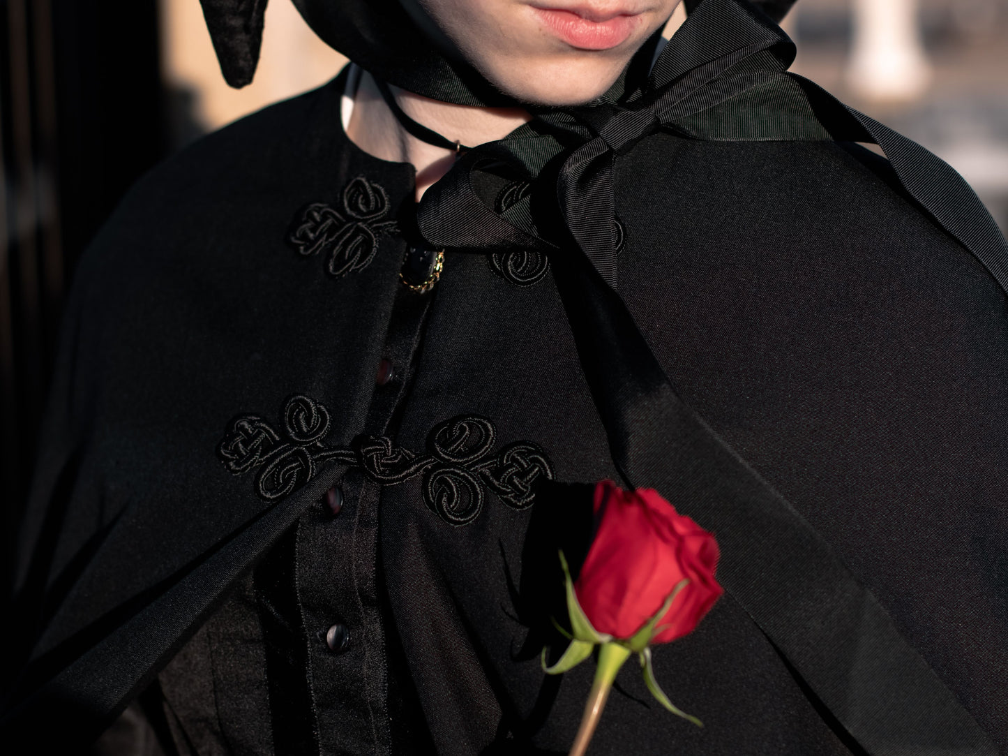 A close-up shows two frog closures on a black wool cape worn by a model. The model holds a red rose.