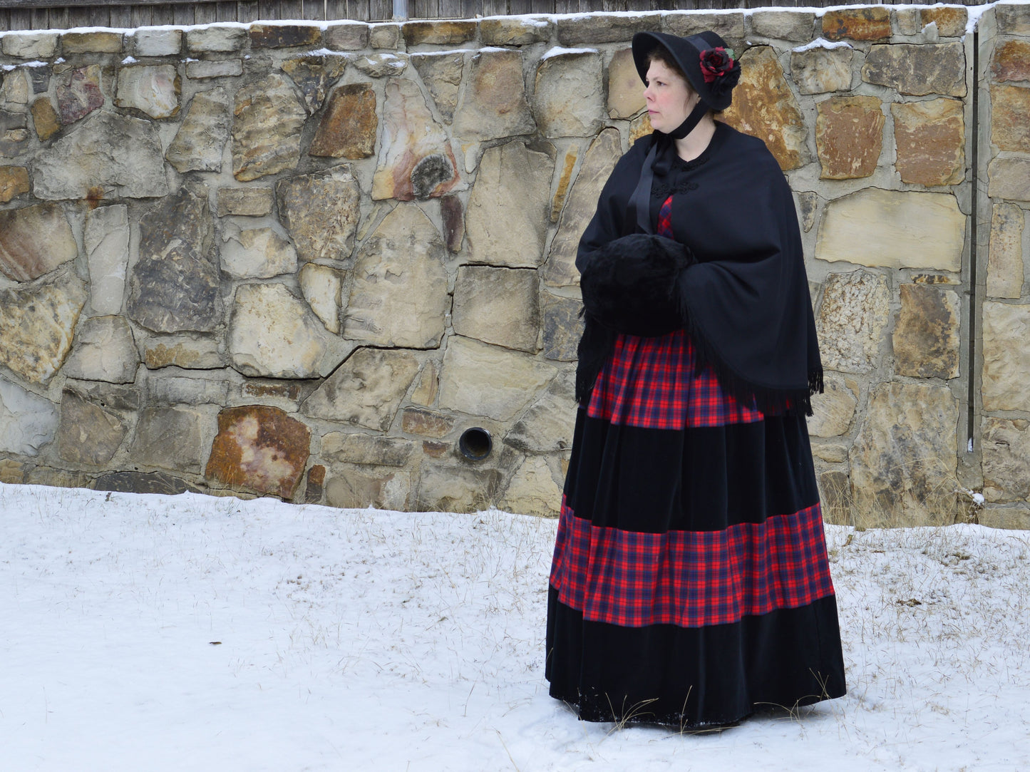 Model stands in the snow against a stone wall. She wears a red plaid 1840s dress with a black wool cape and holds a fur muff