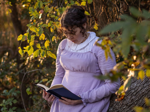 A girl dressed in Regency attire leans against a leafy tree while reading a book