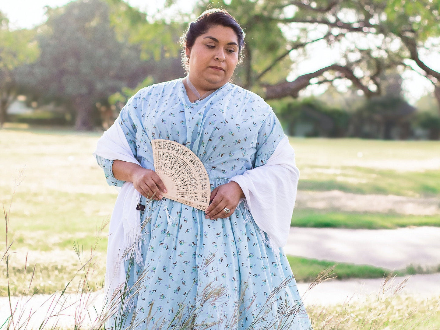 A model wears a blue floral Victorian fan dress with a white shawl. She holds a fan and soft light bathe her and the surrounding trees
