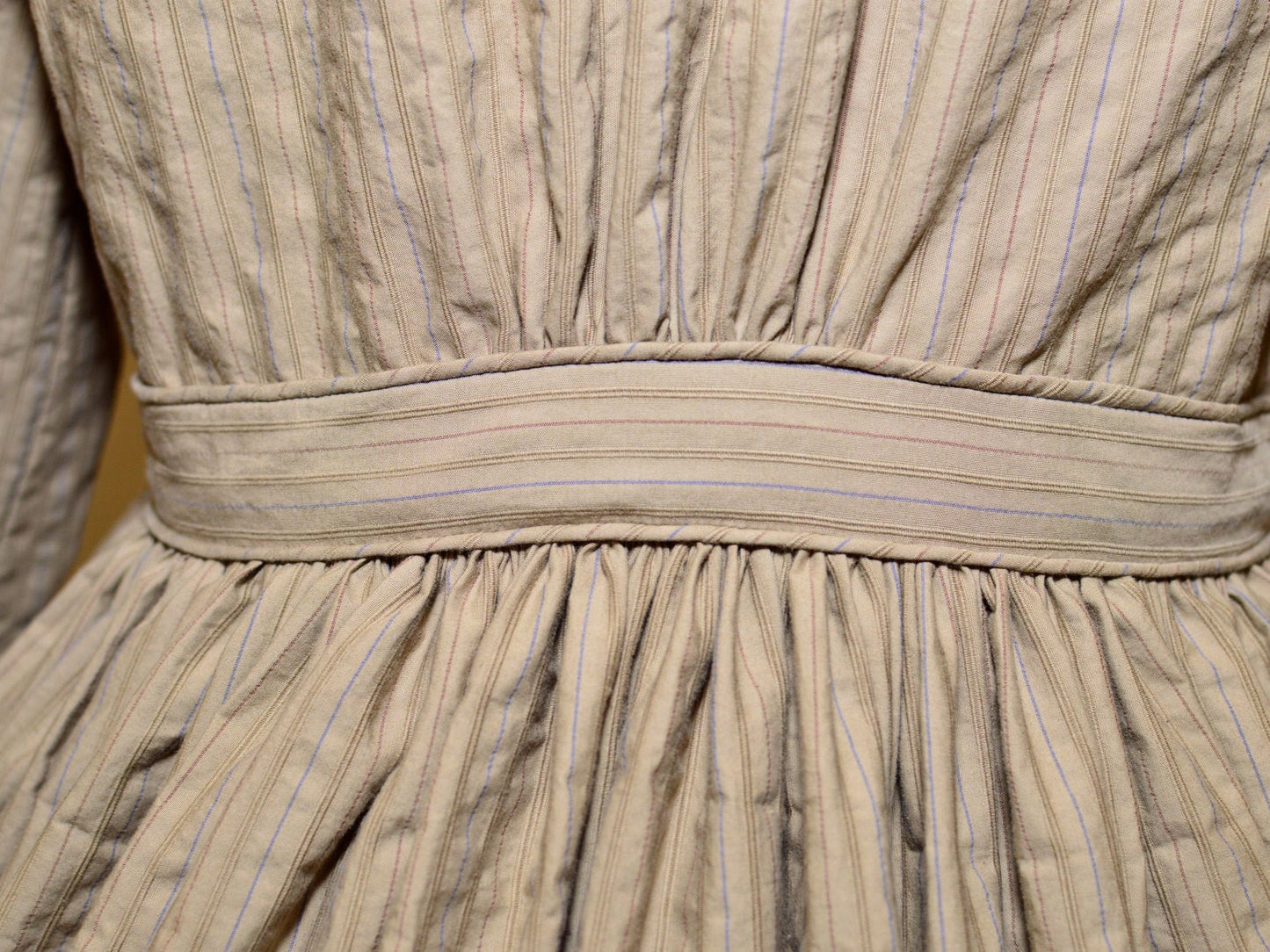 A close-up photo shows the piping on the waistband of a brown 1860s dress