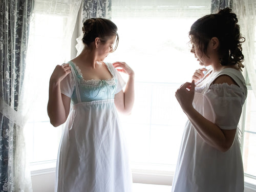 Two girls stand by a bay window while getting dressed in Regency undergarments