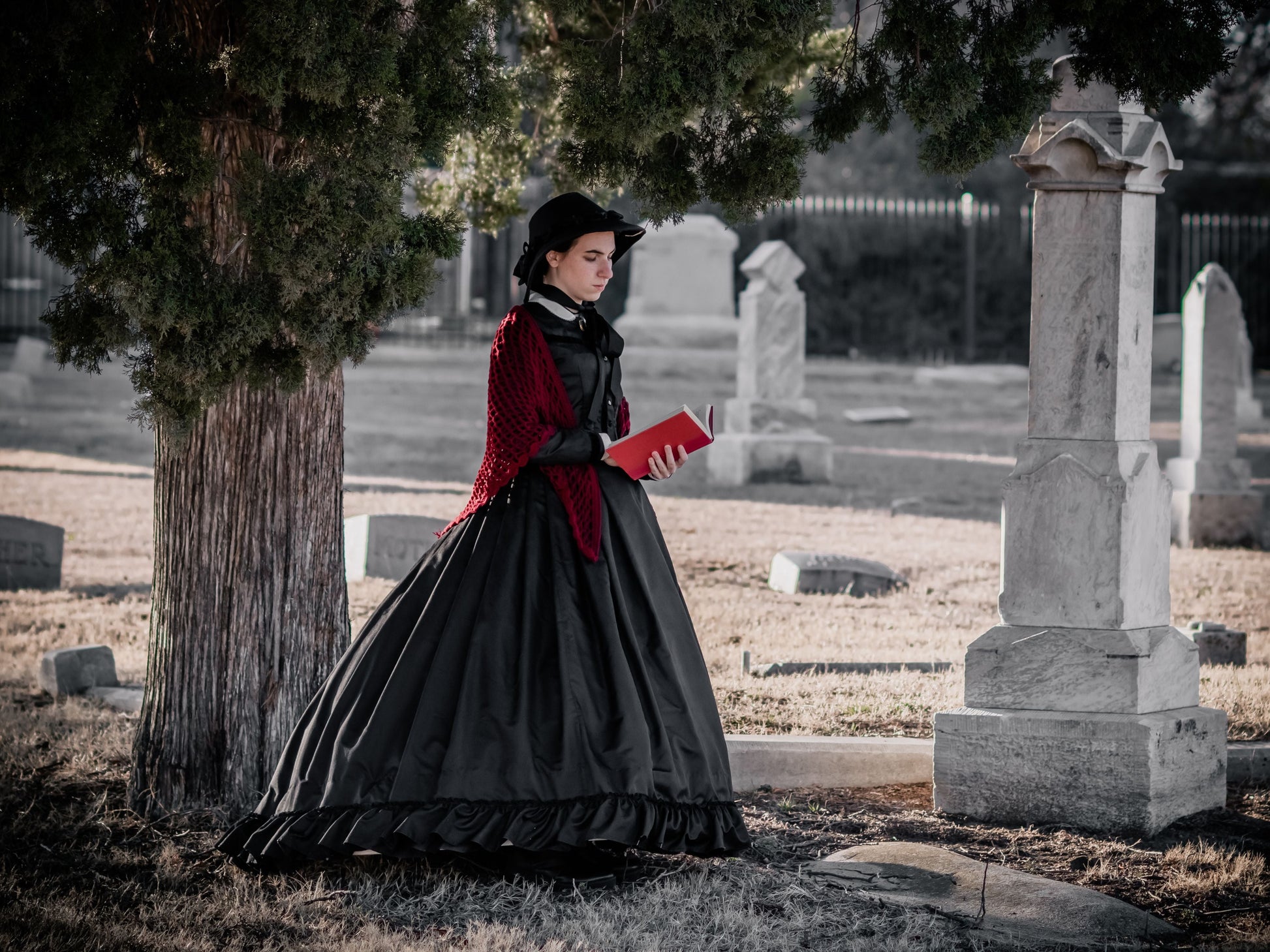 A model dressed in a black silk 1860s dress with a red shawl and red book stands in an old cemetery