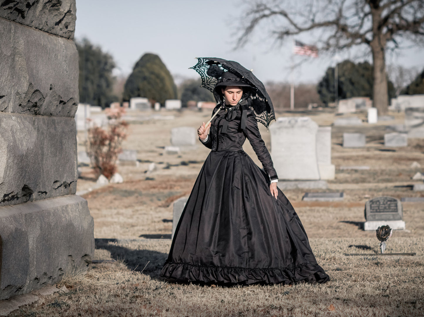 Model wears an all-black silk mourning dress from 1860s and holds a black lace umbrella.  She stands in a cemetery