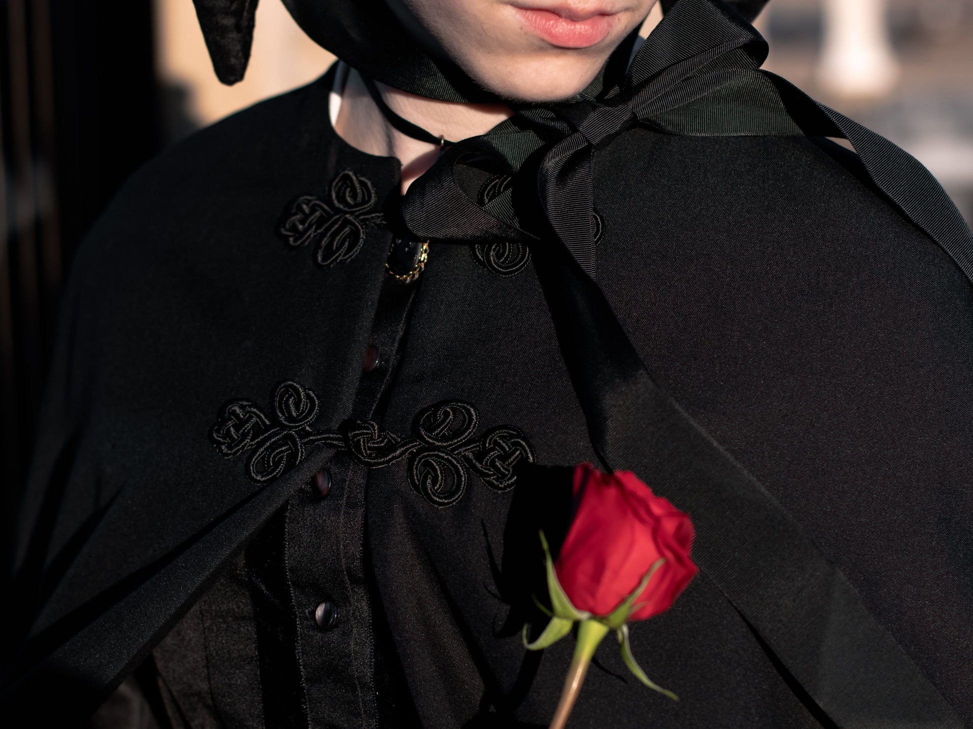 A close-up shows two frog closures on a black wool cape worn by a model.  The model holds a red rose.