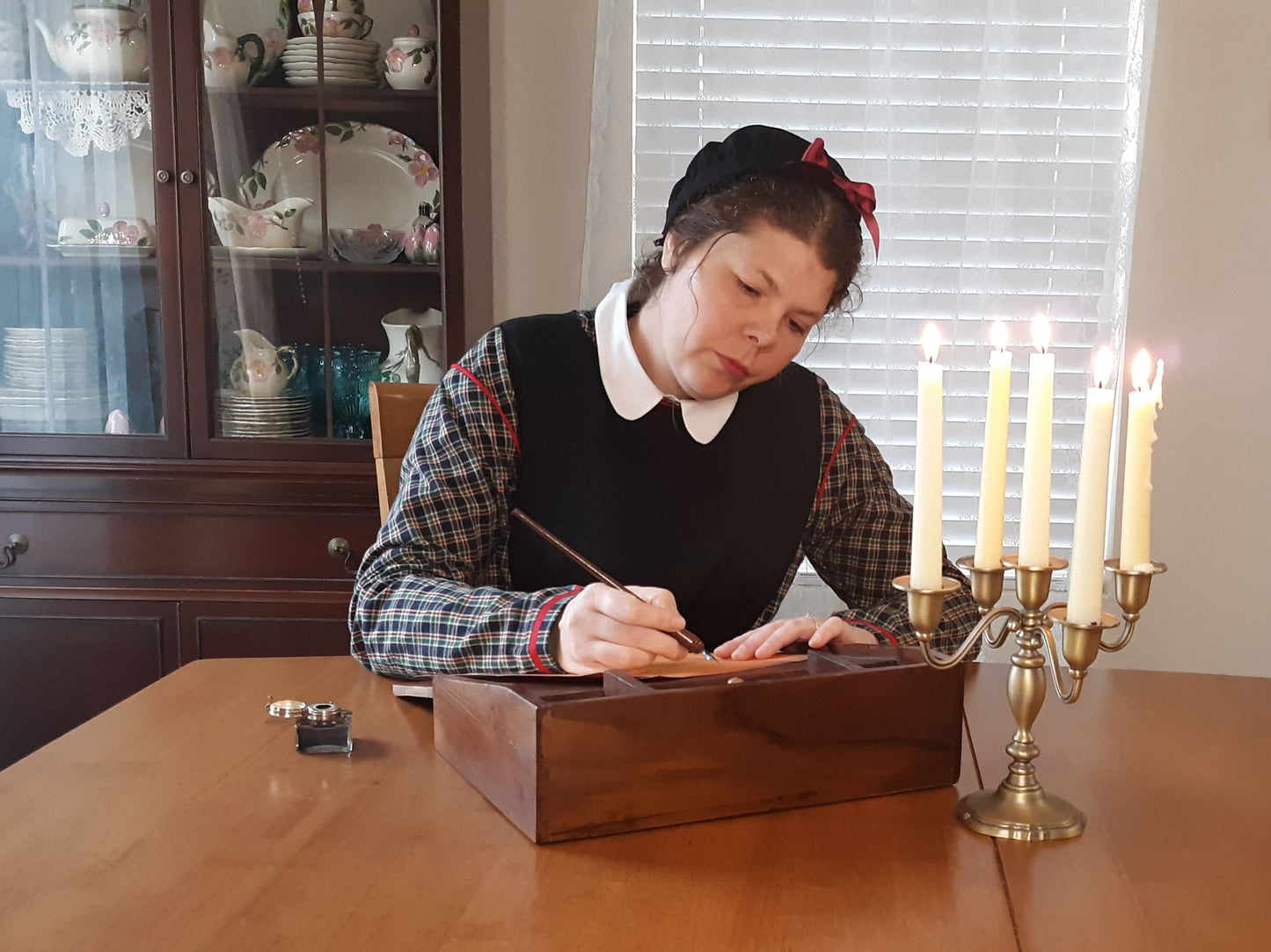 Model sits at a wooden table, writing with a dip pen on a writing desk.  A candelabra burns beside the desk.  The model is wearing an 1860s dress, pinafore and cap.