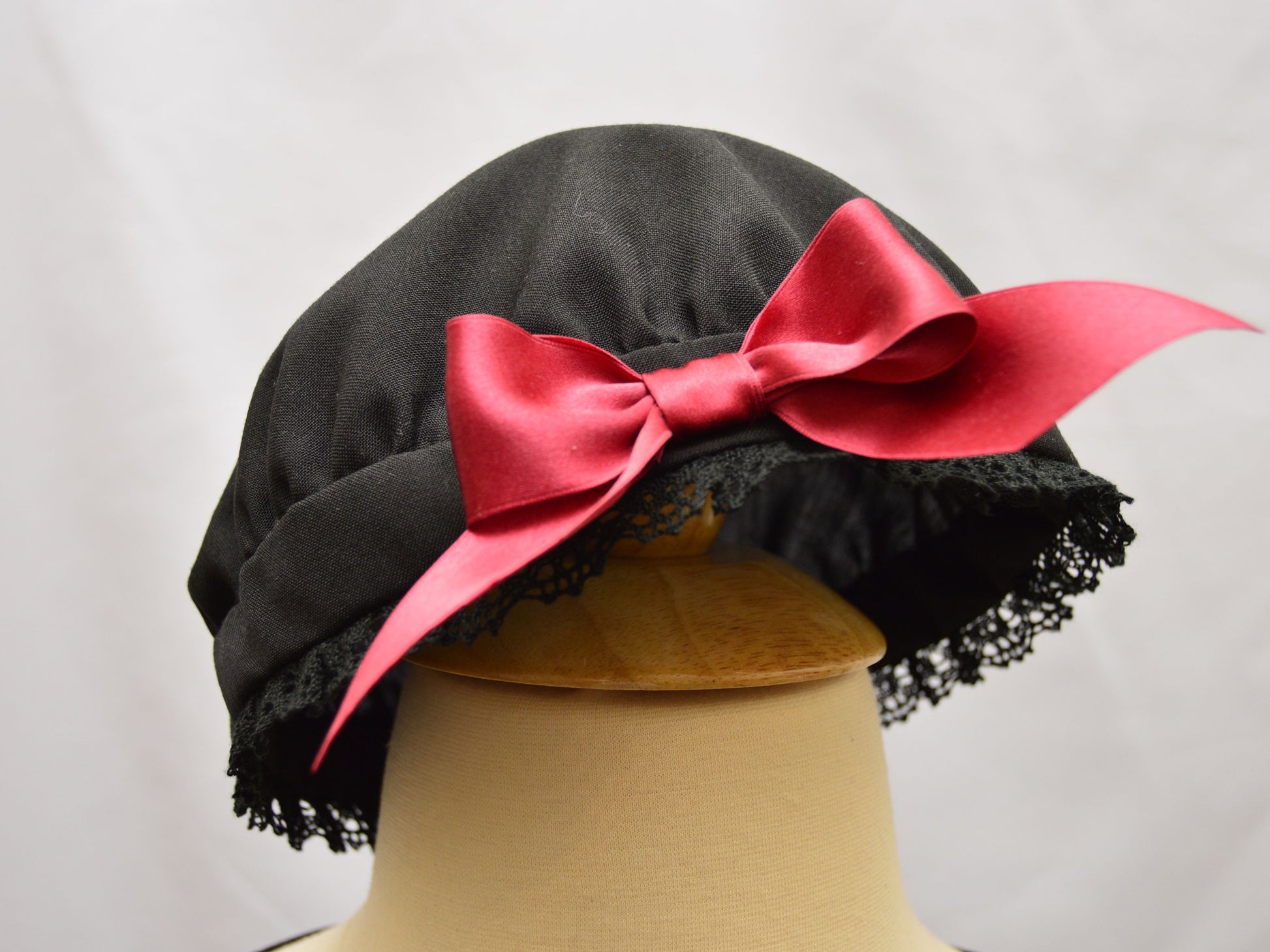 A close-up photo of a black wool cap.  It is adorned with a red silk bow and black cotton lace edging