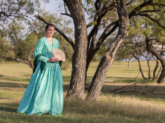 A model wears a mid-Victorian dress and walks outdoors.  She Is holding a fan and wearing a shawl