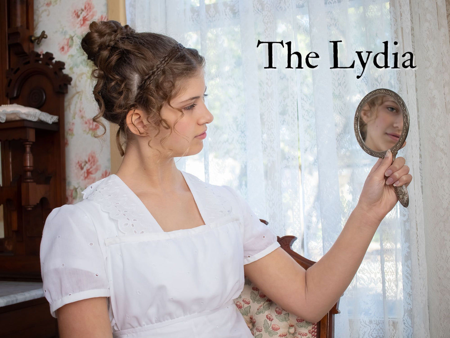 A girl wears all white Regency attire while looking at her reflection in a small hand mirror.