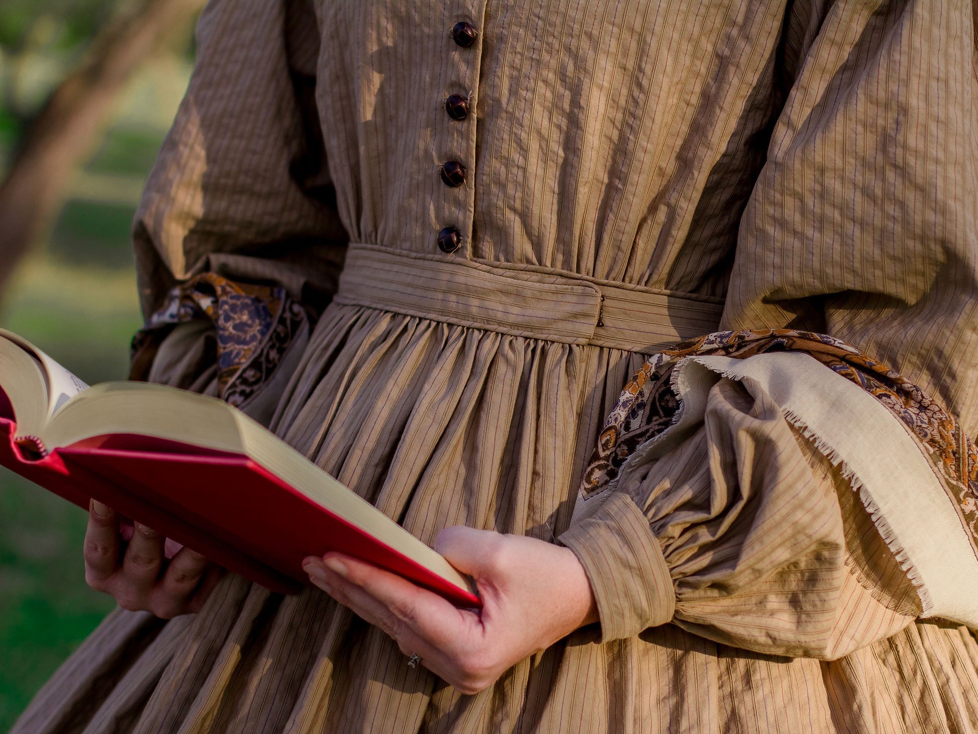 A close-up photo shows the waistband of a model wearing an 1860s dress - clearly illustrating the dog-leg closure.  The model holds a red book in her hands.