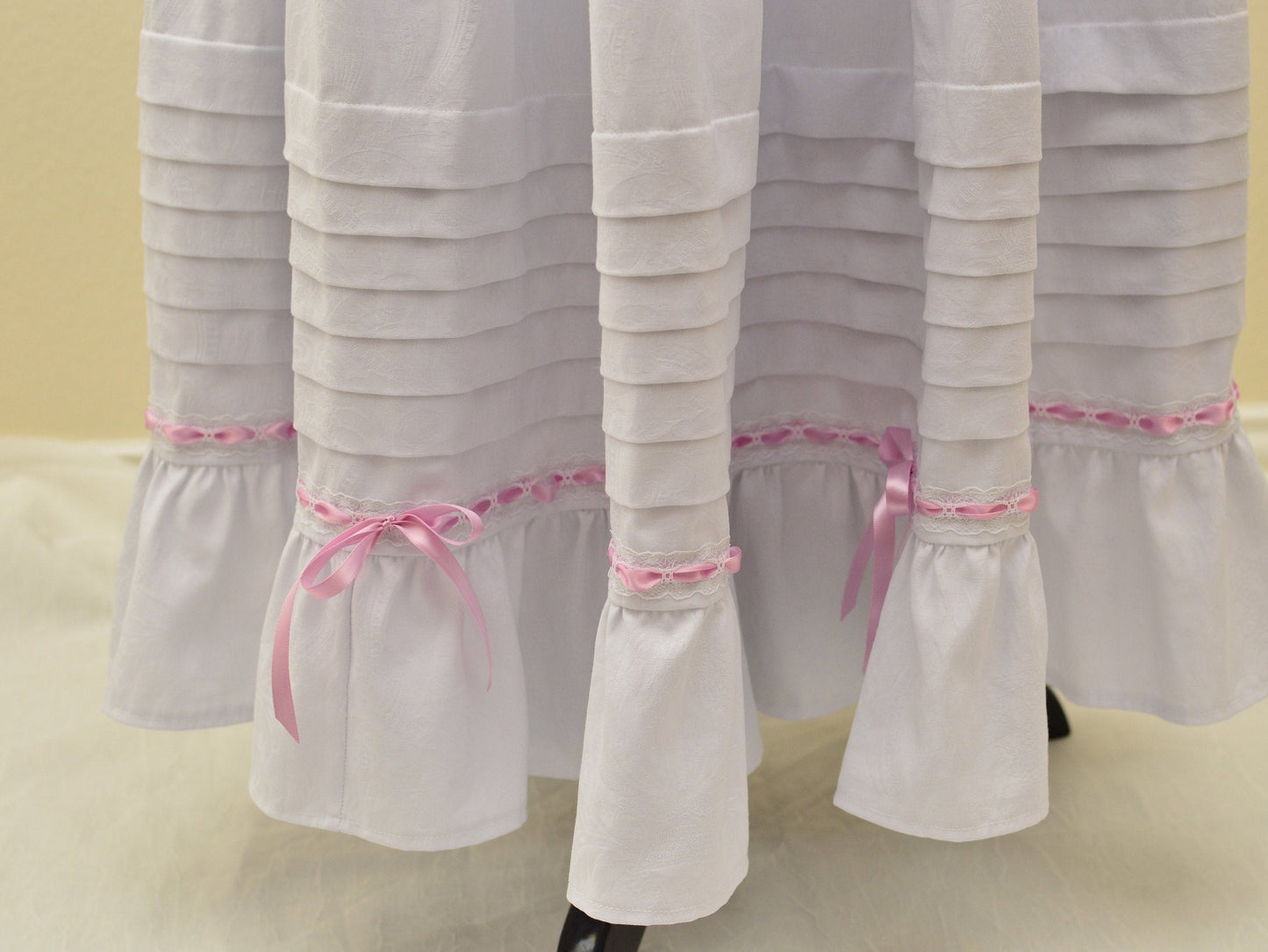 A close-up shows the hem of a Victorian petticoat: six deep tucks, lace beading with pink satin ribbon and bows, and a ruffle